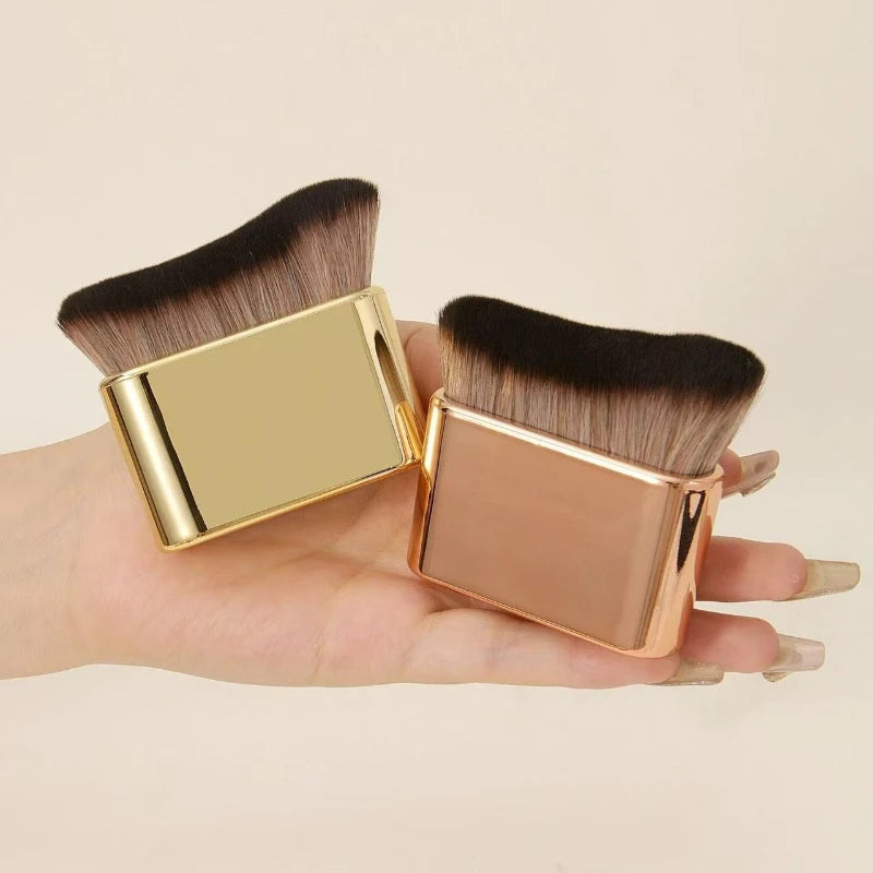 A pair of gold-colored precision hand-held foundation makeup brushes with dark brown bristles are held in a person's hand.