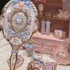 A hand holds a decorative makeup mirror set featuring pink, blue, and gold accents on a white background.