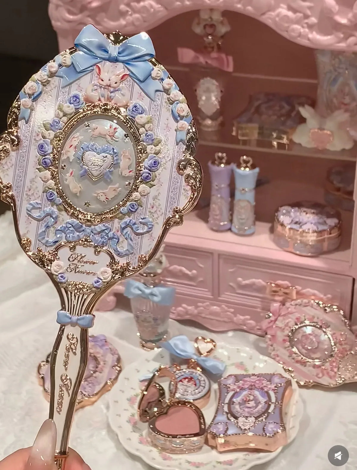 A hand holds a decorative makeup mirror set featuring pink, blue, and gold accents on a white background.
