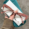 Une paire de lunettes de lecture à œil de chat rose or, posées sur un tissu sarcelle sur un fond gris.