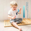A young boy is shown using a blue silicone toothbrush on his teeth while sitting on a wooden mat.