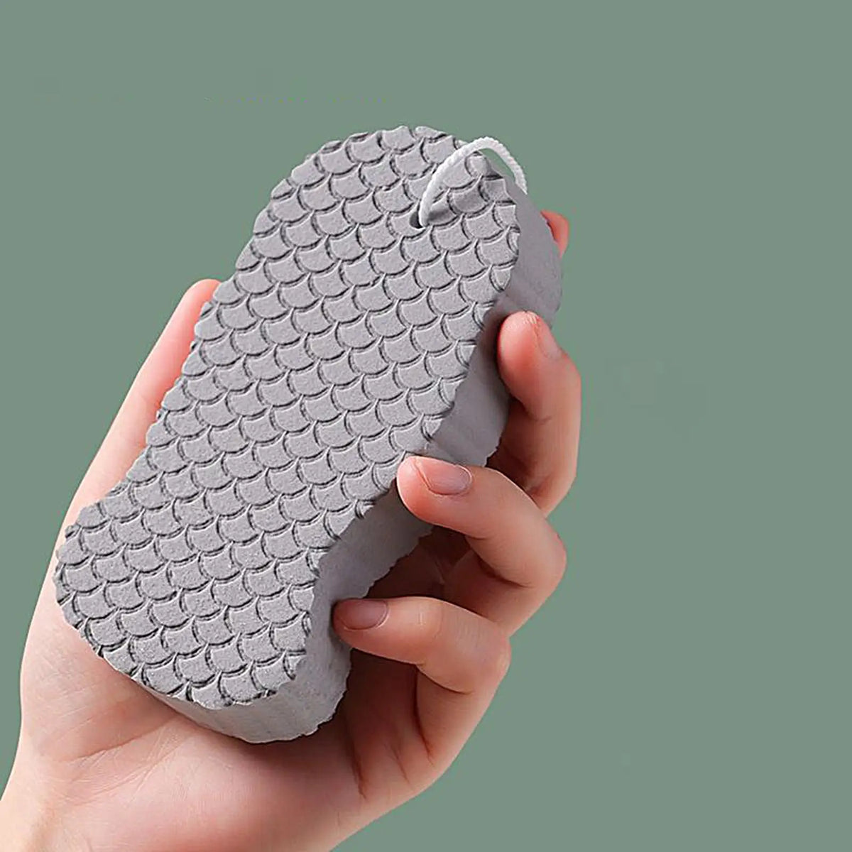 A hand holds a gray exfoliating bath sponge set with a textured, scale-like surface against a muted green background.