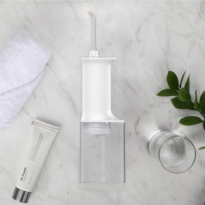 A clear portable dental water flosser device with a narrow nozzle and a glass jar is displayed on a marble surface.