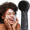 A woman is using a black silicone facial cleansing brush on her face to remove dirt and oil.