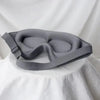 A gray 3D sleeping eye mask is displayed on a white fabric background.