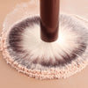 A close-up of a brown makeup brush with light pink powder on the bristles resting on a beige surface.
