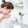 A hand is using a green ultrasonic facial cleansing brush on a sink, with skincare products visible in the background.