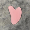 Masajeador facial de cera rosa en forma de corazón sobre una superficie texturizada gris.