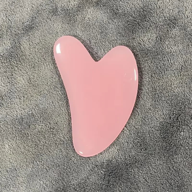 Masajeador facial de cera rosa en forma de corazón sobre una superficie texturizada gris.