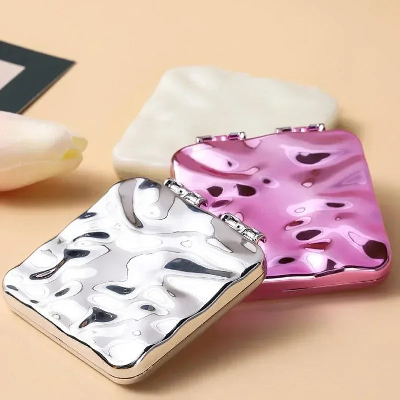 Miroir de maquillage carré pliable en argent et rose marbré, posé sur une surface beige clair.