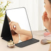 A portable folding makeup mirror with a black stand reflects a person's face.