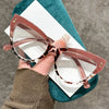 Une paire de lunettes à œil de chat rose gold et tendance posée sur un tissu sarcelle sur un fond gris.
