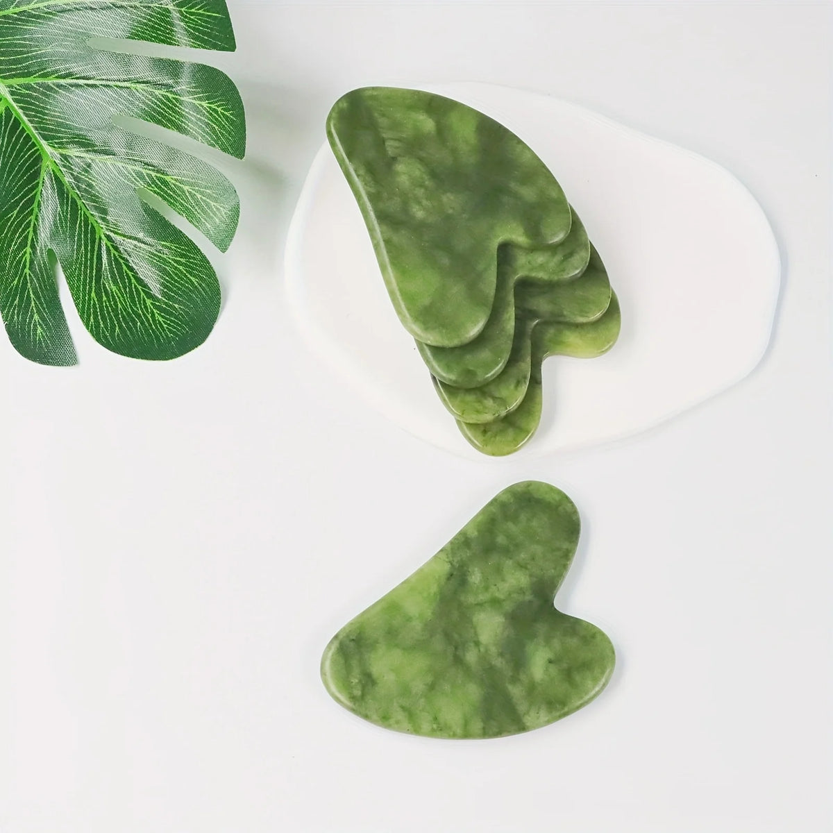 Green jade gua sha tool shaped like a leaf, resting on a white plate.