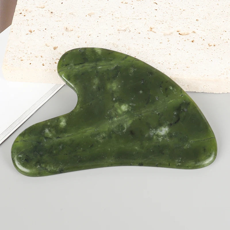 A smooth, oval-shaped green jade gua sha tool resting on a white surface next to a block of light-colored stone.