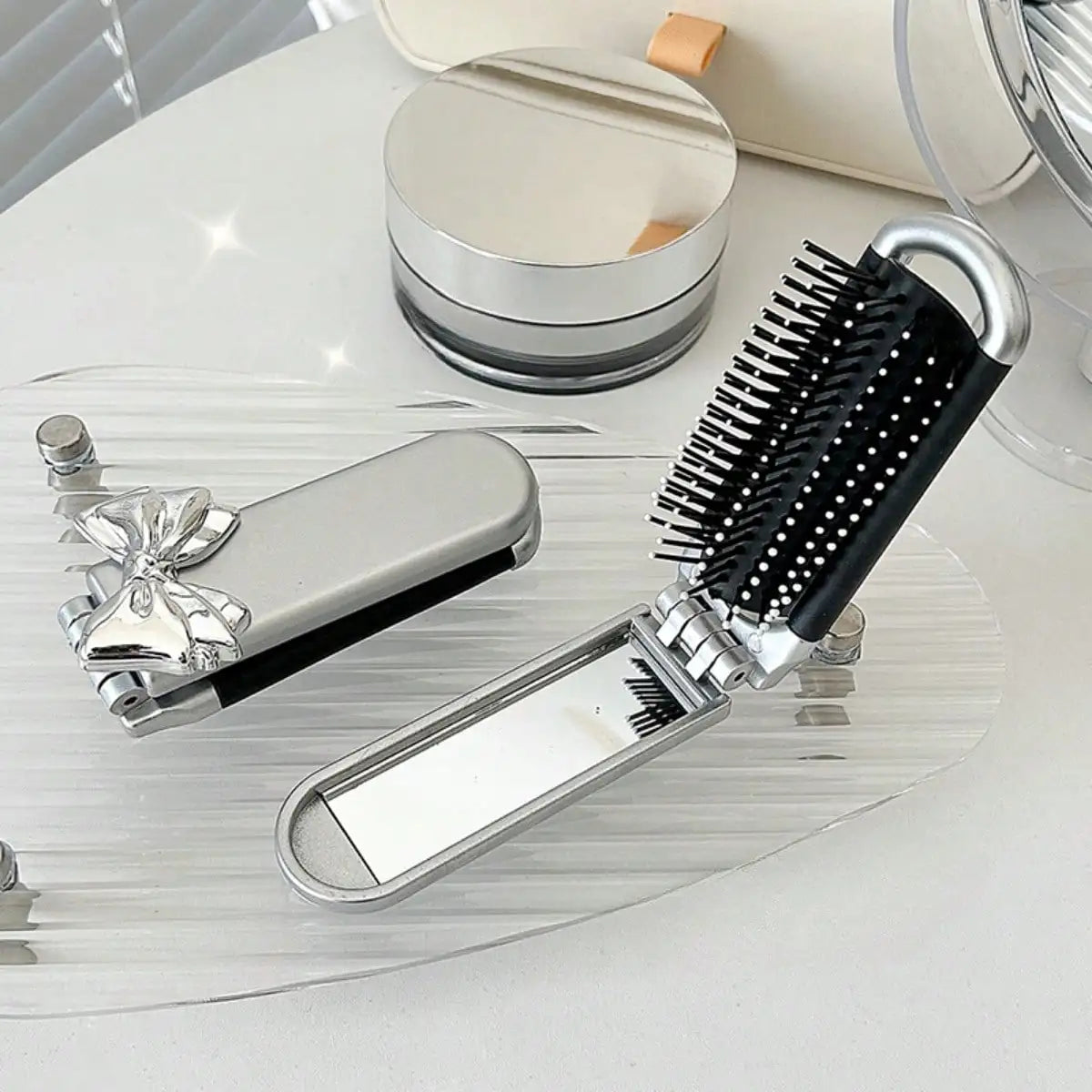 A silver portable compact comb with an attached mirror and a black bristled comb is displayed on a clear glass surface.