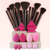 A collection of makeup brushes and sponges in various shades of pink and black arranged on a light wooden box.