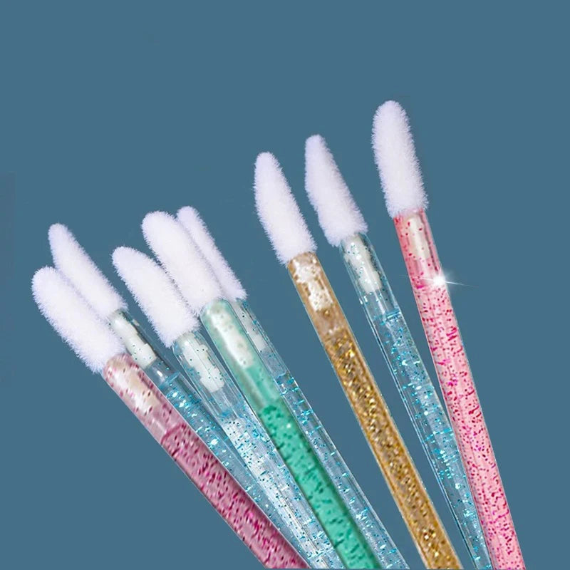 A collection of crystal lip brushes and lash spoolies in various shades of pink, blue, and gold against a dark background.