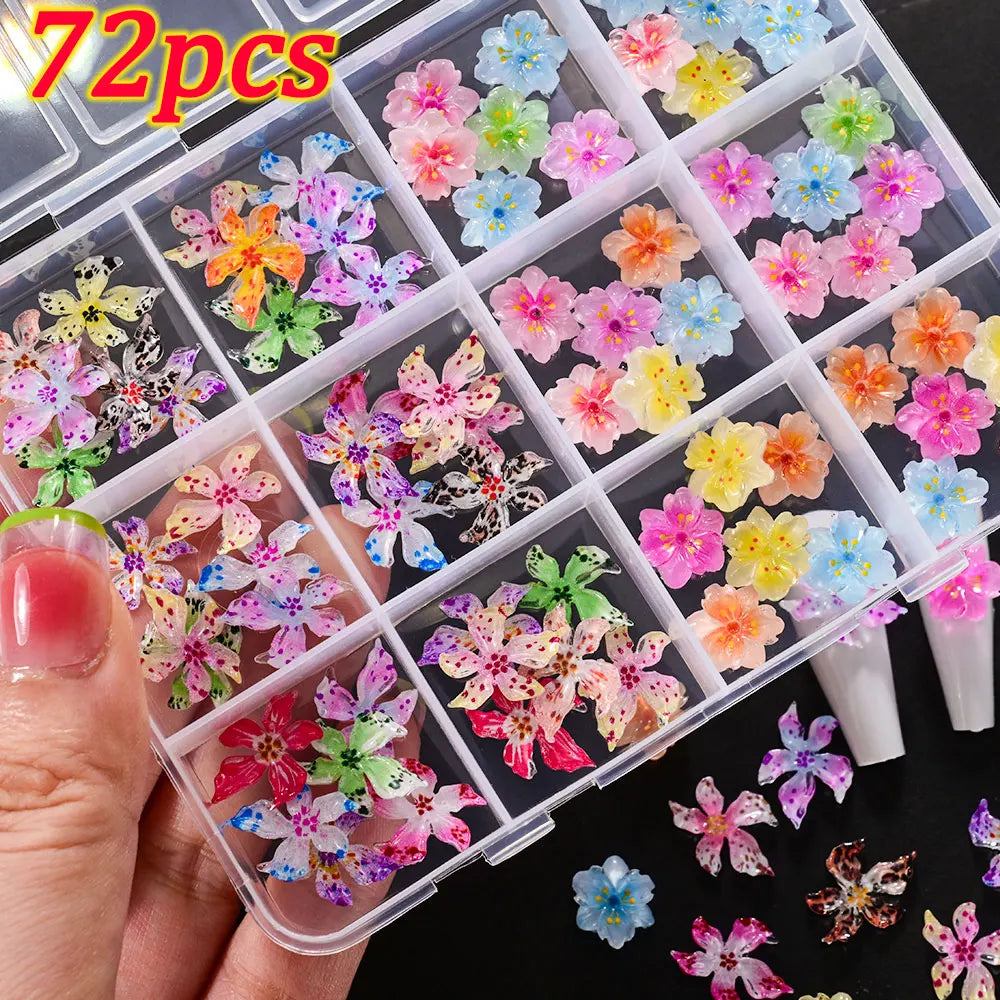 A hand holds an open clear plastic box filled with colorful floral resin nail charms featuring hibiscus flowers in various sh