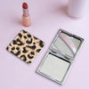 A portable makeup mirror with a leopard print cover and a lipstick tube on a pink surface.