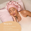 A woman wearing a pink satin sleep cap and top rests on pillows, demonstrating hair curling results.
