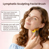 A woman is using a yellow and brown lymphatic sculpting facial brush on her face.