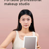 A woman holds a portable professional makeup studio LED mirror with touch dimming on a light pink background.