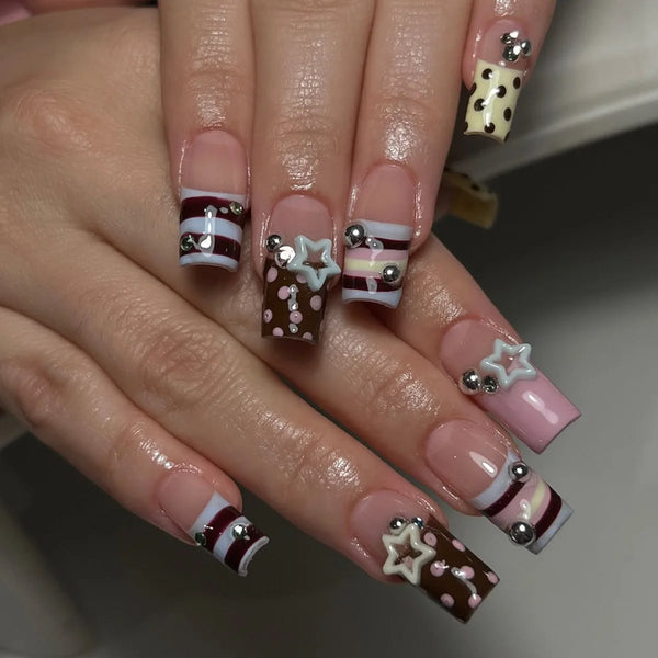 A hand displays press-on nails featuring a French wave design with stars and polka dots in brown, white, and pink tones.