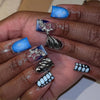 A hand displays a set of nude pink press-on nails featuring leopard print and 3D silver charms on the tips.