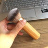 A hand holds a bamboo airbrush foundation and bronzer brush resting on a wooden desk next to a silver laptop.