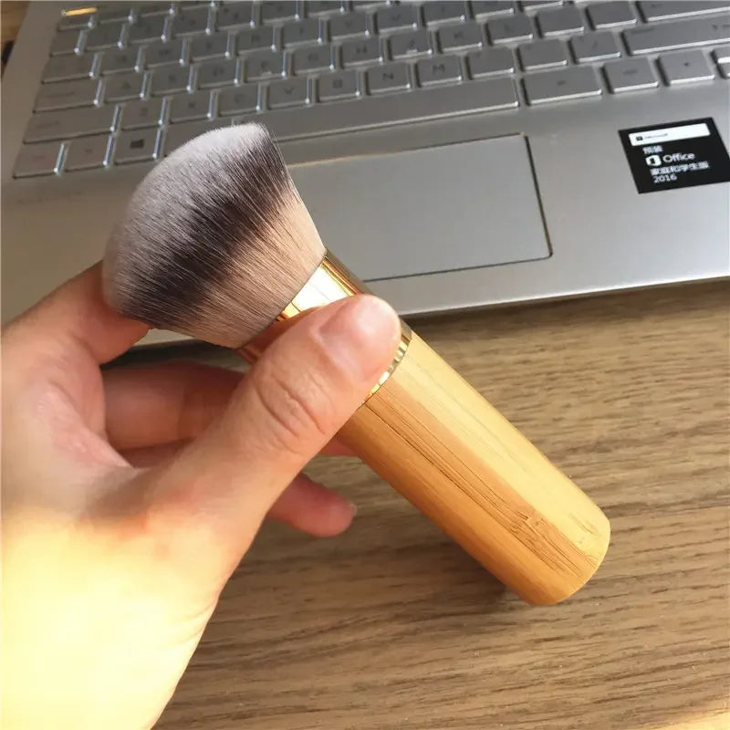 A hand holds a bamboo airbrush foundation and bronzer brush resting on a wooden desk next to a silver laptop.