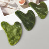 A green jade gua sha tool is shown on a light surface next to a piece of fruit.