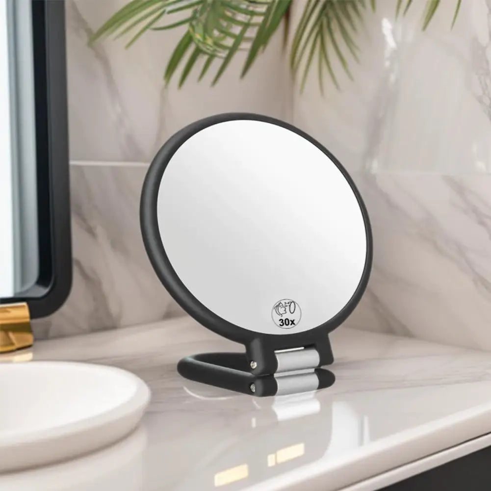Un miroir grossissant rond noir avec une poignée argentée repose sur un comptoir blanc.