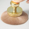 Un grand pinceau de maquillage en poudre avec une poignée dorée et des poils roses clairs est tenu par un doigt.