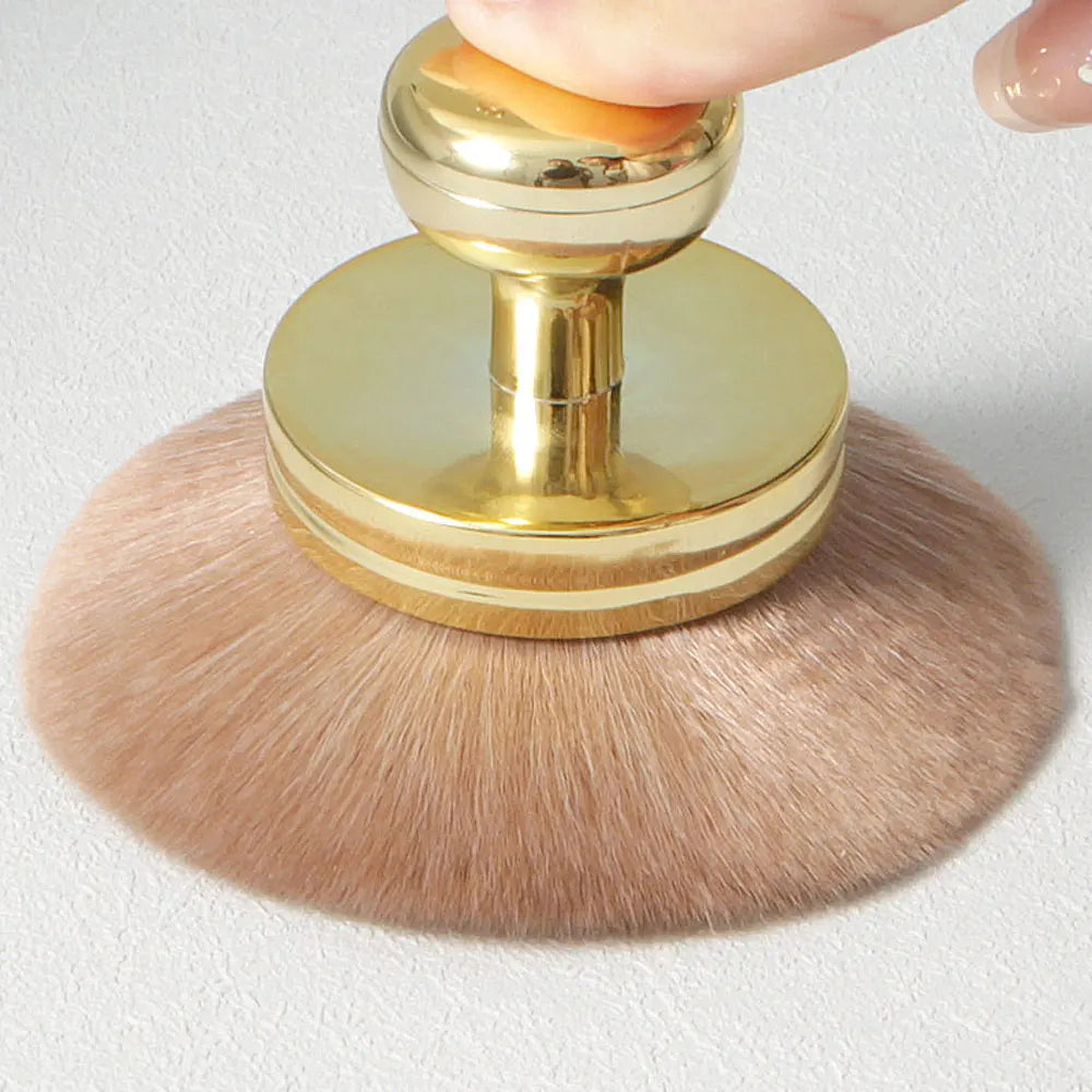 Un grand pinceau de maquillage en poudre avec une poignée dorée et des poils roses clairs est tenu par un doigt.