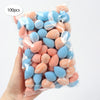A hand holds a clear plastic bag filled with 100 reusable expandable makeup sponges in various pastel pink and blue colors.