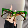 A hand holds a pair of green tortoiseshell reading glasses with clear frames against a light background.