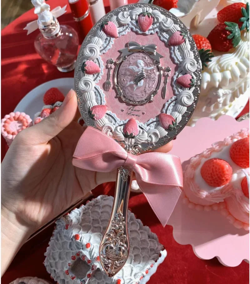 A hand holds a silver makeup mirror set decorated with pink strawberries and a pink ribbon on a red background.