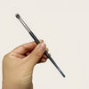 A hand holds a black precision concealer brush with a silver ferrule and a fine tip.