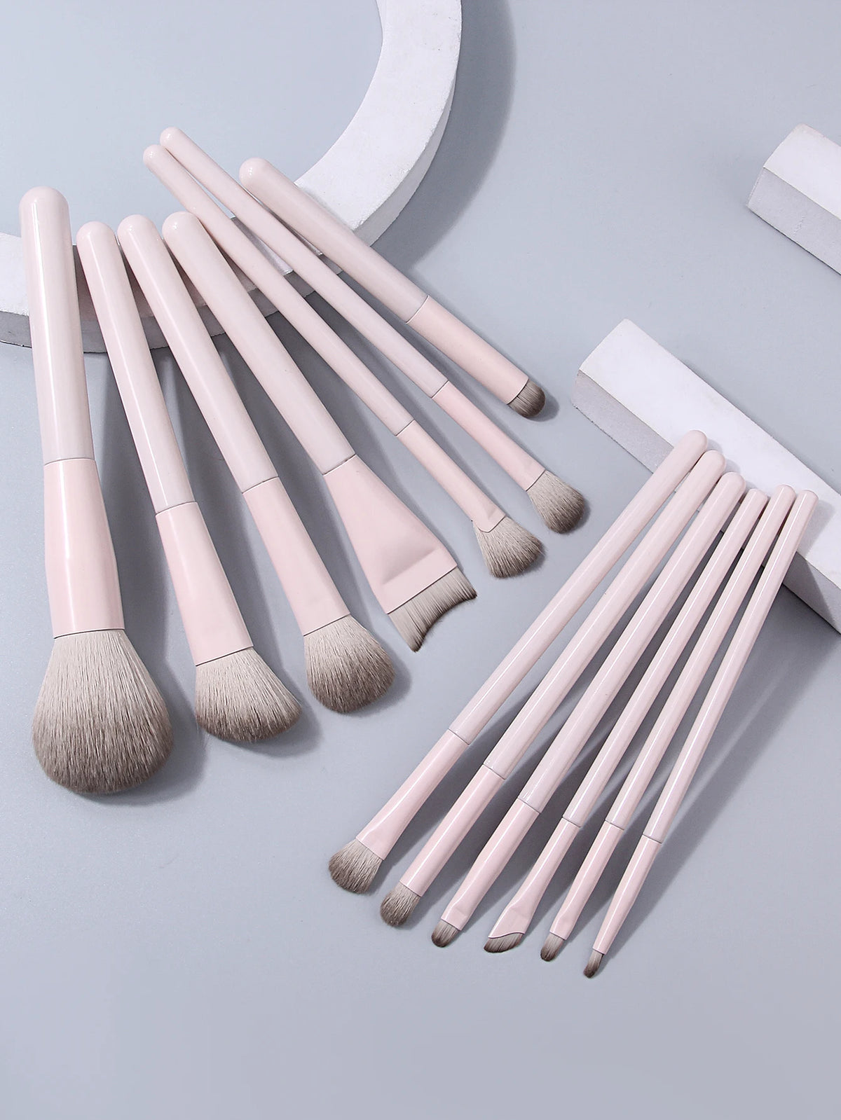 A set of thirteen professional makeup brushes in a pale pink color arranged on a light gray surface.