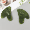 A green jade gua sha tool is shown on a light gray surface next to a white paper, with a portion of a sliced citrus fruit vis