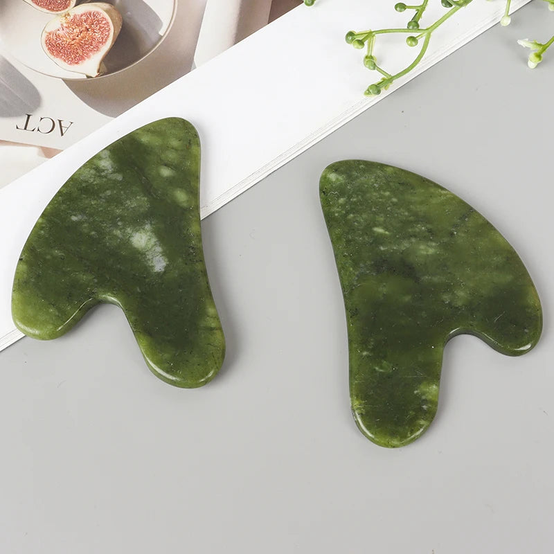 A green jade gua sha tool is shown on a light gray surface next to a white paper, with a portion of a sliced citrus fruit vis