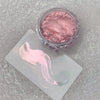 A jar of pink glitter powder with a pink cosmetic brush stroke displayed on a white card.