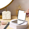 Une main tient un miroir de maquillage carré pliable posé sur un support blanc à côté d'un tulipe rose.