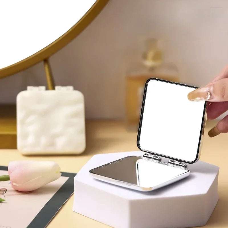 Une main tient un miroir de maquillage carré pliable posé sur un support blanc à côté d'un tulipe rose.