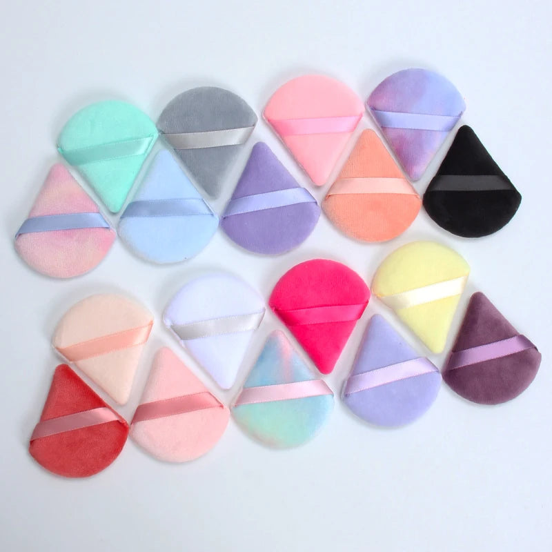 Un ensemble de six éponges de maquillage triangulaires aux couleurs pastel et foncées, disposées sur une surface blanche.