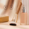A gold-toned angled contoured makeup foundation brush next to its bottle and vegan cruelty-free certification logos.