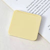 A square, pale yellow double-sided portable makeup mirror resting on a white surface next to a piece of paper.