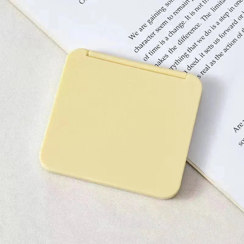 A square, pale yellow double-sided portable makeup mirror resting on a white surface next to a piece of paper.