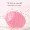 A pink silicone facial cleansing brush with textured surfaces resting on white exfoliating beads.