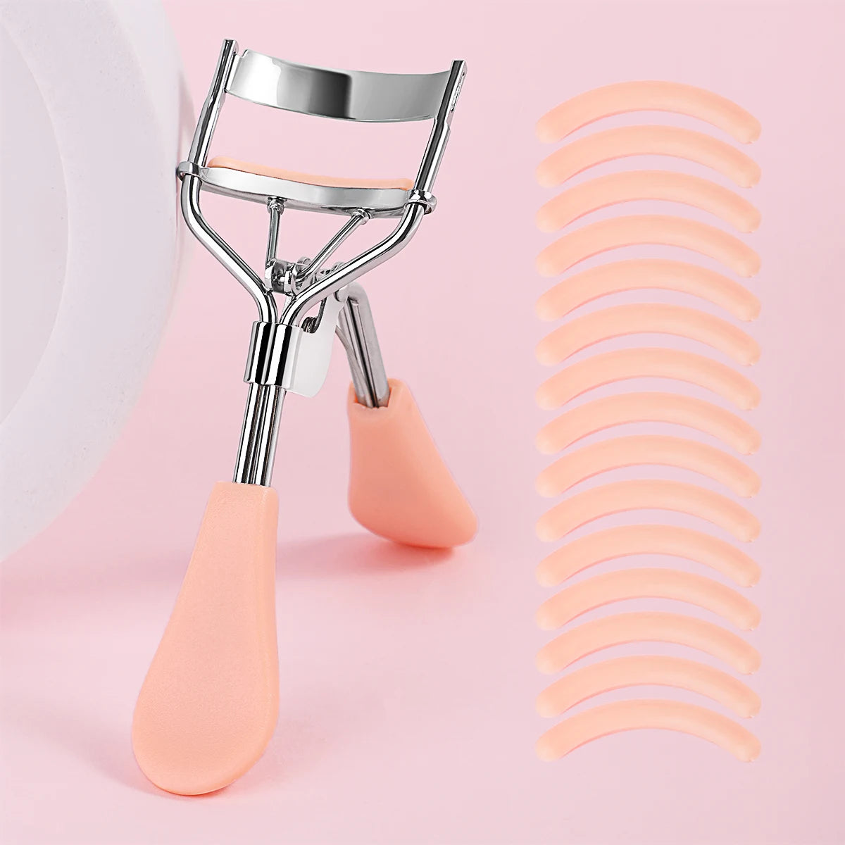 A professional stainless steel eyelash curler set with pink silicone applicators shown against a light pink background.